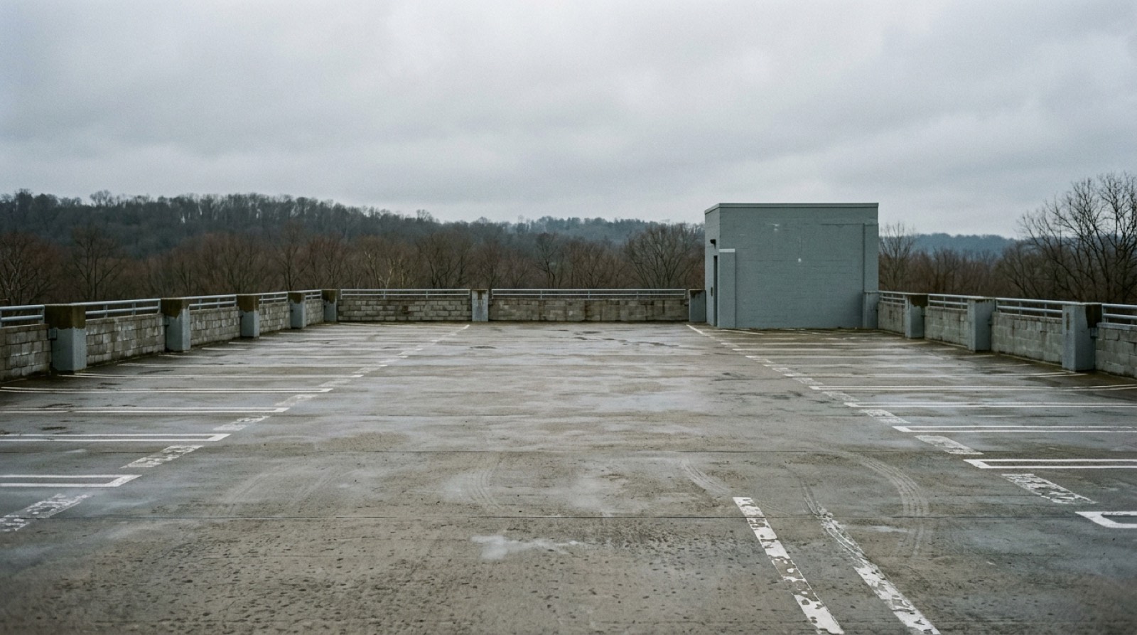 The open upper floor of a concrete parking structure, empty, with painted lines and a low perimeter wall, under an overcast sky